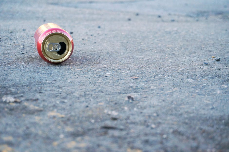 A discarded soda can lying on a rough, asphalt surface. The can is made of aluminum with a metallic finish, featuring pink and gold coloring around the top rim and body. The pull-tab is open, exposing